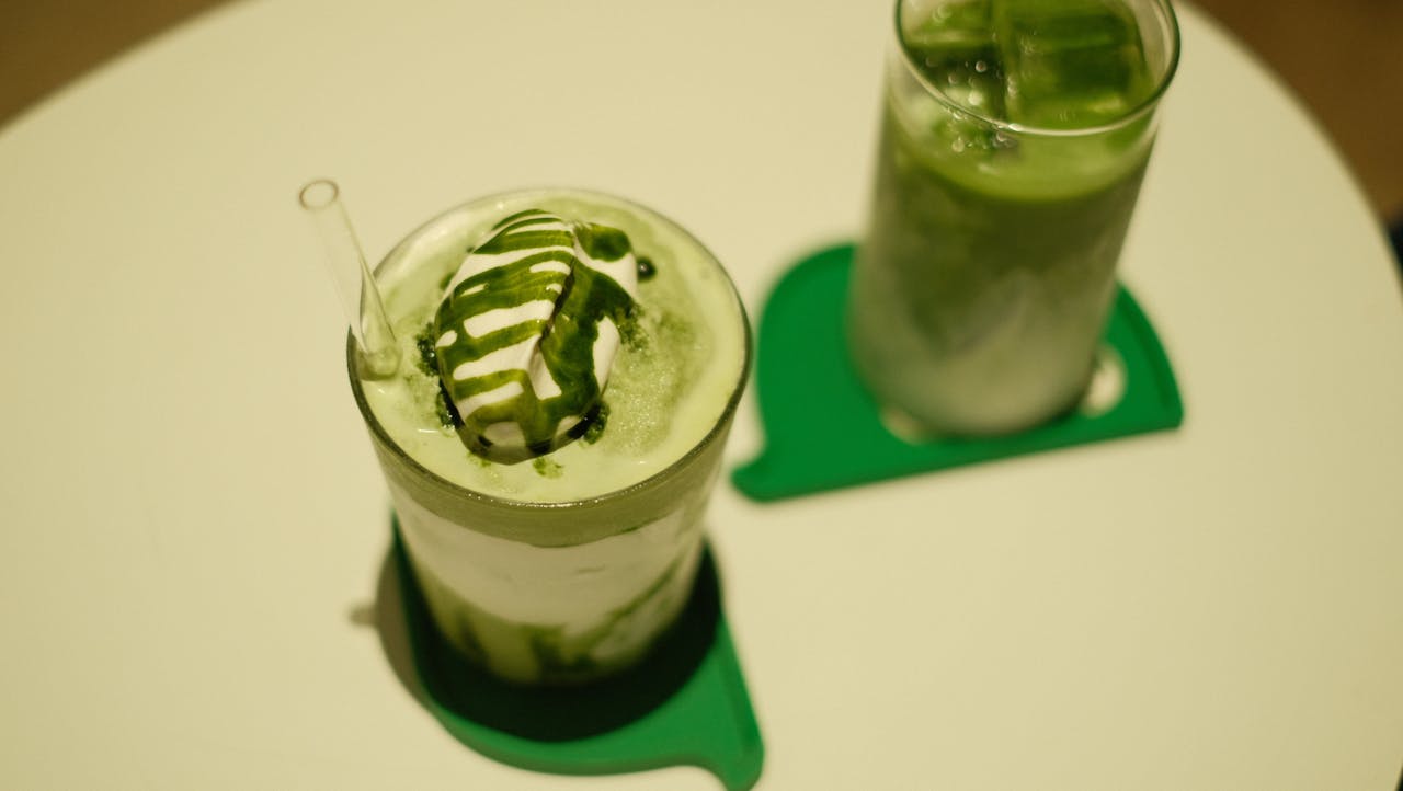 Two iced matcha lattes topped with cream and matcha drizzle on a table.